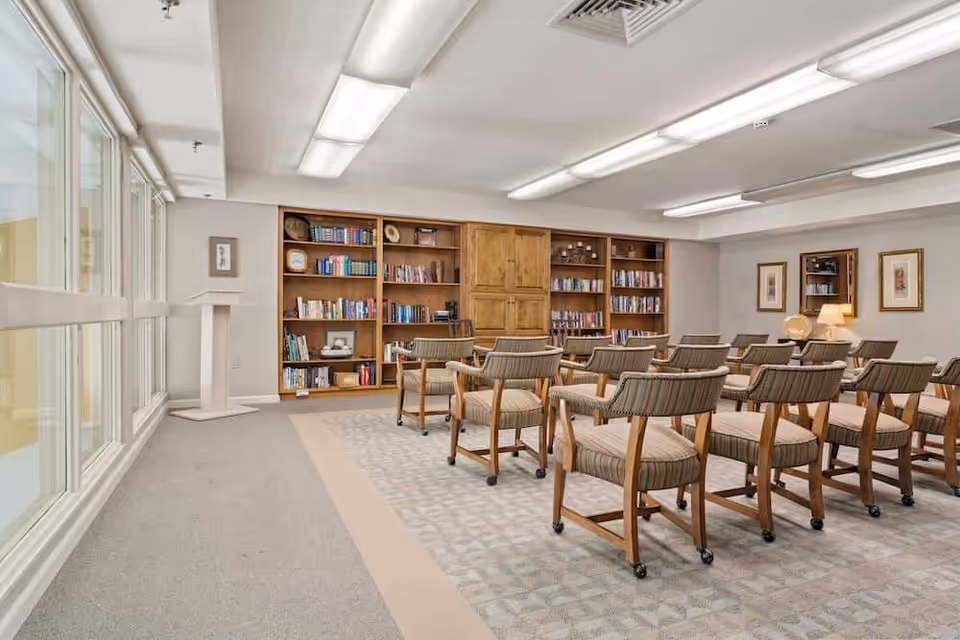 A bright community room with rows of upholstered chairs facing bookshelves and large windows.