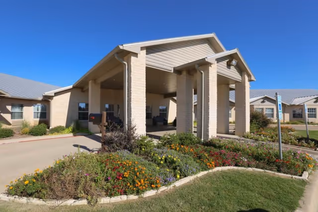 Exterior view of Stillhouse Rehabilitation And Healthcare Center showing the entrance with a covered drop-off area supported by stone pillars, surrounded by a landscaped garden with colorful flowers and a clear blue sky.