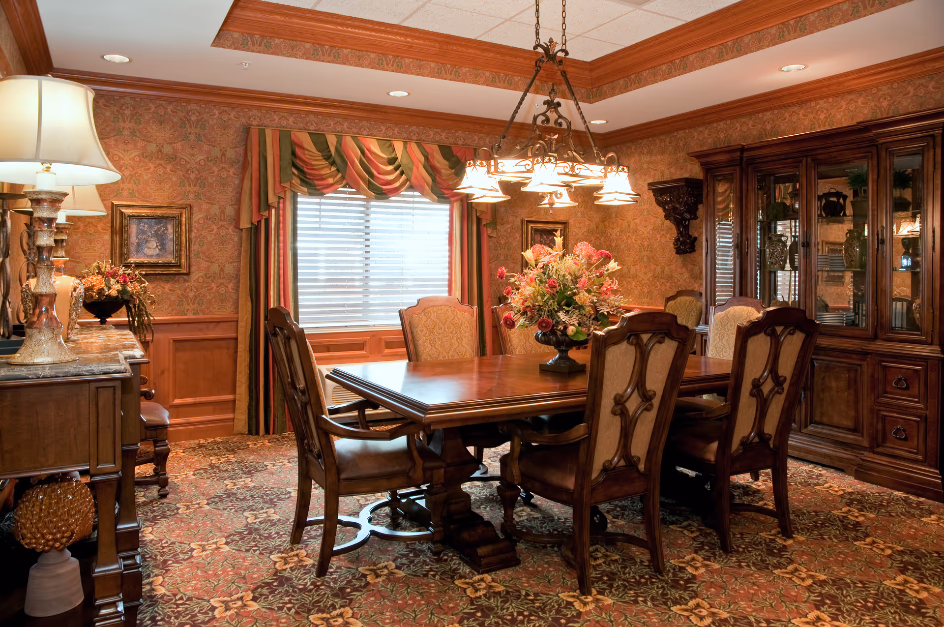 A traditional dining room with a wooden dining table surrounded by six upholstered chairs. The room features ornate wallpaper, a large window with striped curtains, a chandelier hanging above the table, a floral centerpiece on the table, a wooden cabinet with glass doors displaying decorative items, and a sideboard with lamps and floral arrangements.