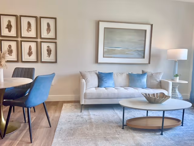 A modern living room with a light beige sofa adorned with three blue cushions, a round white coffee table with a decorative bowl, a white side table with a lamp, and a round dining table with blue and gray chairs. The walls are decorated with framed artwork featuring feathers and a landscape painting.