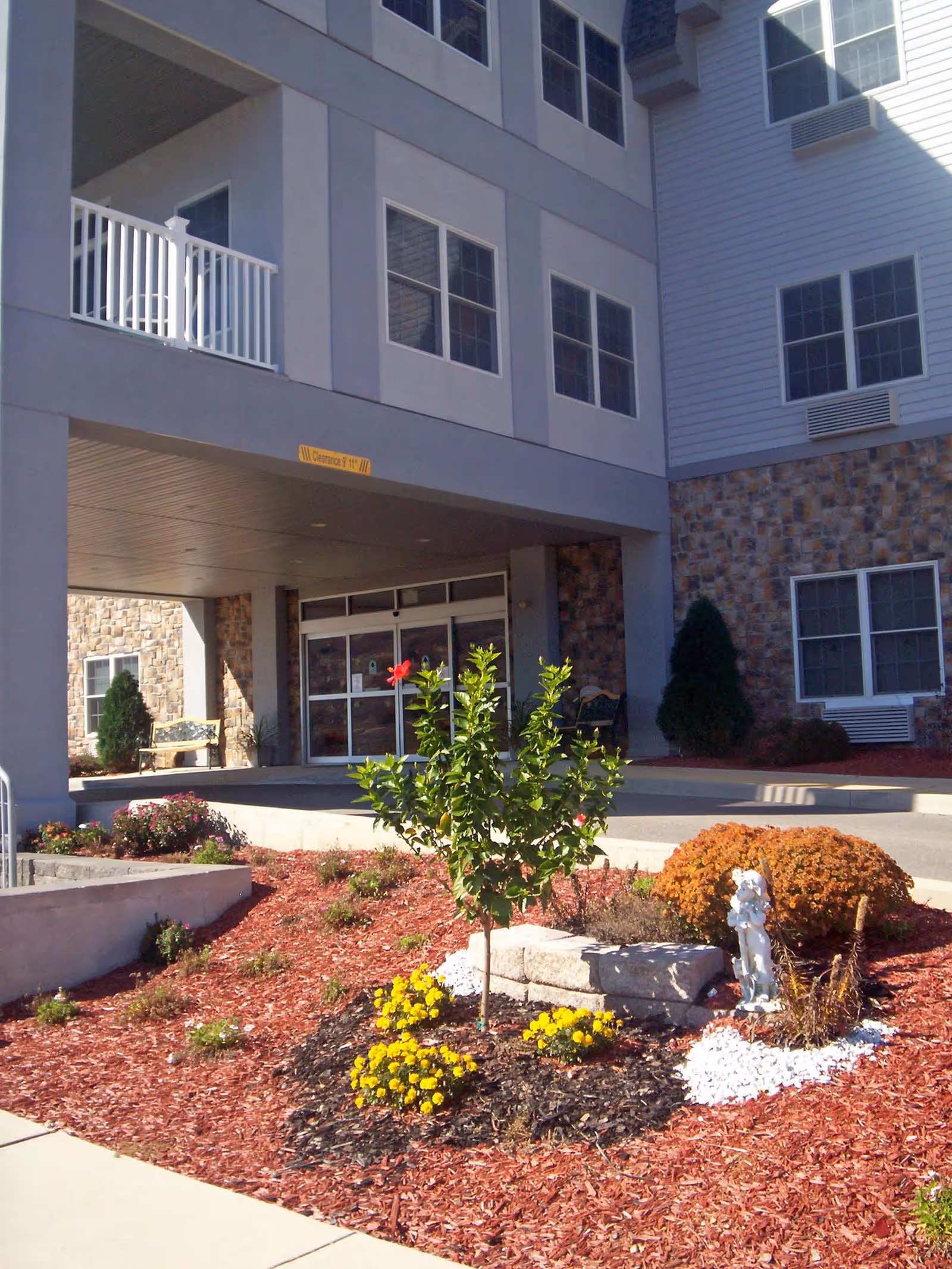 Entrance of The Palmerton facility showing a covered drop-off area with glass doors, surrounded by a landscaped garden featuring a small tree, yellow flowers, a bush, and a small white statue. The building exterior includes stone and siding with multiple windows.
