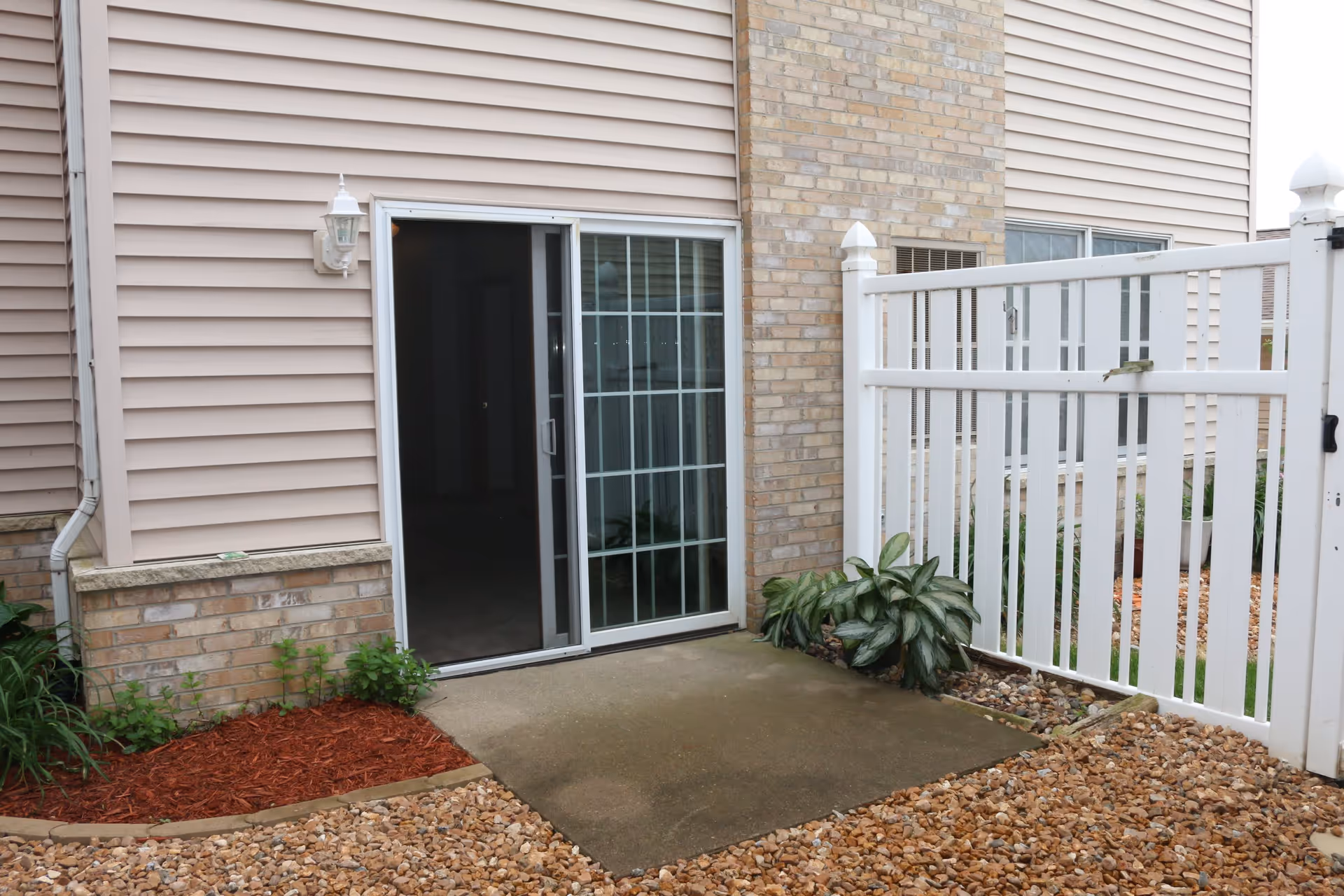 Outdoor patio area with a sliding glass door leading inside. The patio has a concrete floor surrounded by small rocks and mulch with some green plants. There is a white fence on the right side and beige siding with brick on the building exterior.
