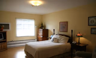 A cozy bedroom with a double bed covered in a cream-colored quilt, two pillows, and a stuffed animal. The room has light yellow walls, a window with blinds letting in natural light, a wooden dresser with a flower arrangement, a nightstand with a decorative lamp, and framed pictures on the walls.