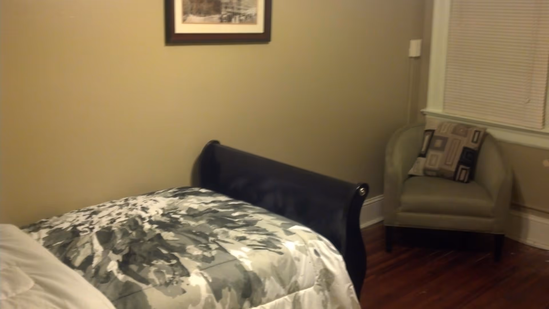 A bedroom with a bed covered in a gray and white patterned comforter, a green armchair with a decorative pillow, a window with closed blinds, and a framed picture on the beige wall.