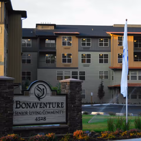 Exterior view of Bonaventure Senior Living Community building with a stone sign in the foreground displaying the facility name and address 4528, surrounded by landscaping and a flagpole with a white flag.