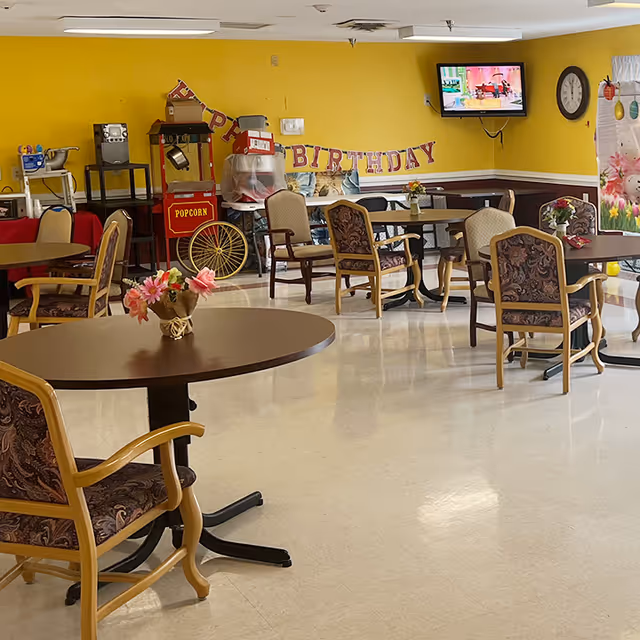 A bright communal dining/activity room with round tables and chairs, floral centerpieces, a popcorn cart, and a 'Happy Birthday' banner on a yellow wall.