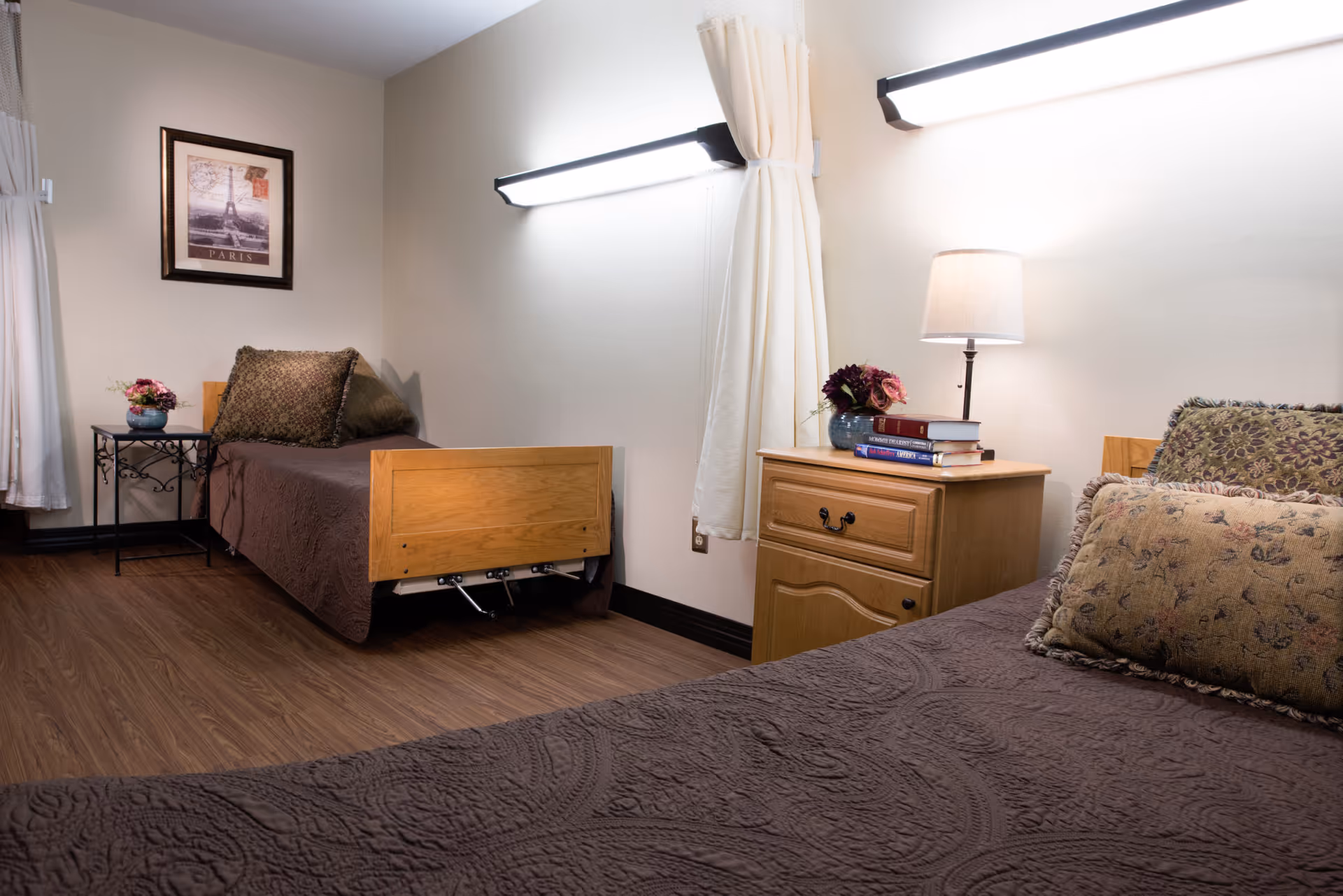 A senior living bedroom with two twin beds, wood nightstand with a lamp and books, decorative pillows, and wall-mounted lights.