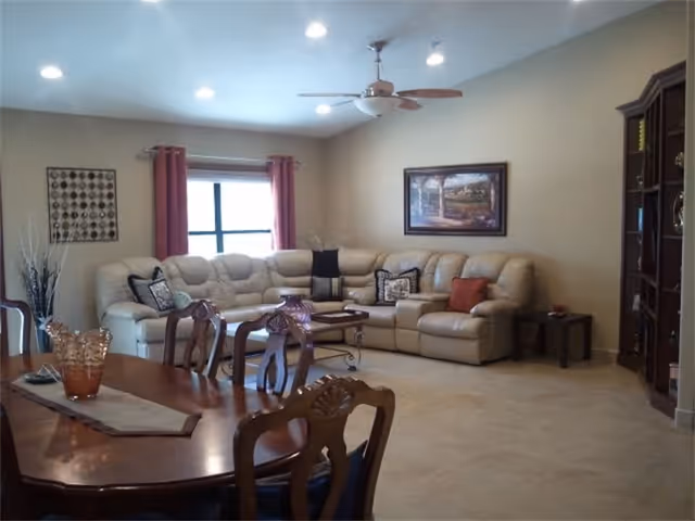 A spacious living room with a large beige sectional sofa adorned with decorative pillows, a wooden coffee table, and a wooden dining table with chairs in the foreground. The room has beige walls, a ceiling fan with lights, recessed ceiling lights, a window with red curtains, and framed artwork on the walls.