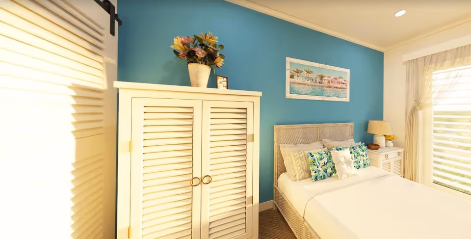 A bright and cozy bedroom with a white bed featuring multiple decorative pillows, a white wardrobe with a flower vase on top, a nightstand with a lamp and small decor, a large window with sheer curtains, and a blue accent wall with a framed beach-themed painting.