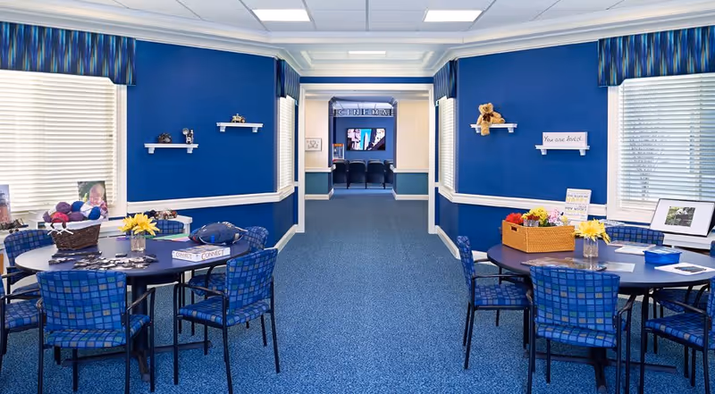 A bright, blue-themed common area with two round tables surrounded by blue checkered chairs. The tables have various items including flowers, a basket with yarn, and papers. The walls are painted blue with white trim and small shelves holding decorative items. At the end of the hallway, there is a room labeled 'CINEMA' with theater-style seating and a large screen.