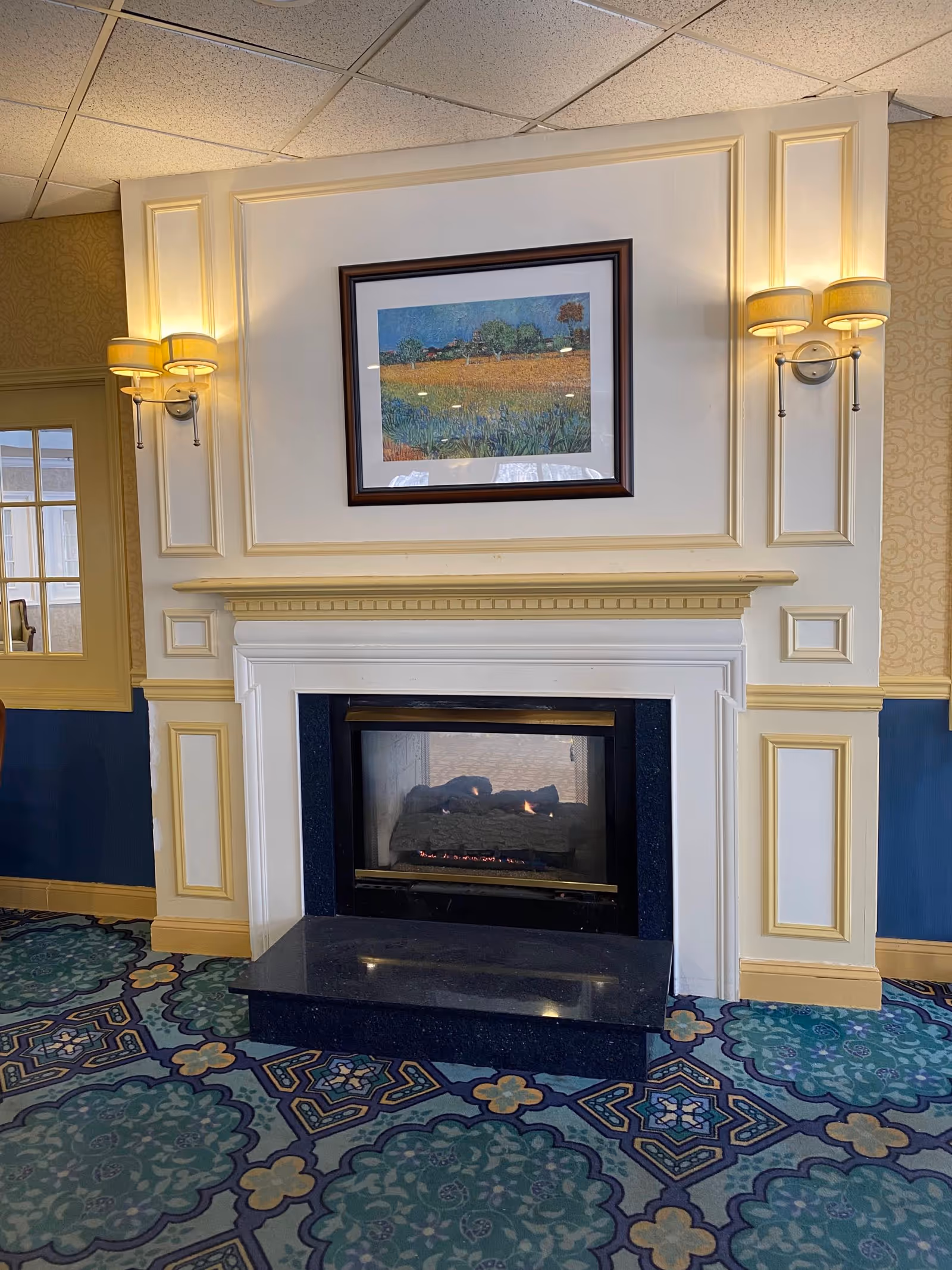 Ornate indoor fireplace with framed artwork above and wall sconces, set in a carpeted common area.
