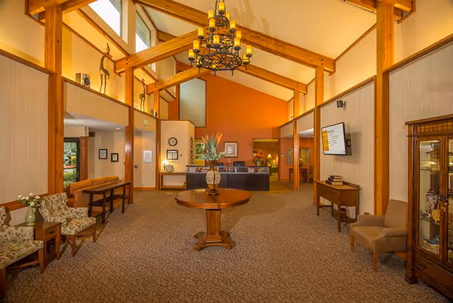 A spacious and warmly lit interior lobby area with high wooden beam ceilings and a large chandelier. The room features a central round wooden table with a decorative vase and flowers, seating areas with upholstered chairs and small tables, a reception desk in the background, and a wall-mounted television on the right side.