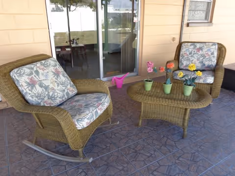 Two wicker rocking chairs and a wicker coffee table with potted flowers on a tiled covered patio in front of a sliding glass door.