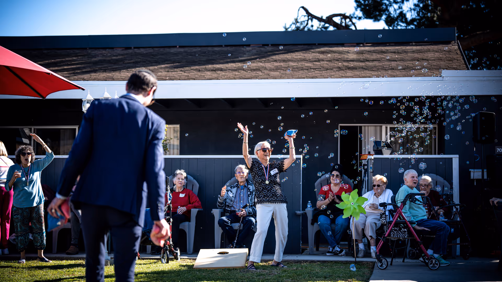 A group of elderly people enjoying an outdoor activity at a senior living facility. One woman in the center is standing with her arms raised, smiling, surrounded by bubbles. Other residents are seated nearby, some clapping and watching the activity. A man in a suit is in the foreground, partially turned away from the camera. The setting is outside with a building in the background and a red umbrella visible on the left.