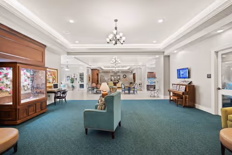 A spacious and well-lit common area in a senior living facility featuring green carpet, comfortable seating including armchairs and sofas, a chandelier, a piano against the wall, and a display cabinet with floral decorations. The area opens into a dining space with tables and chairs visible in the background.