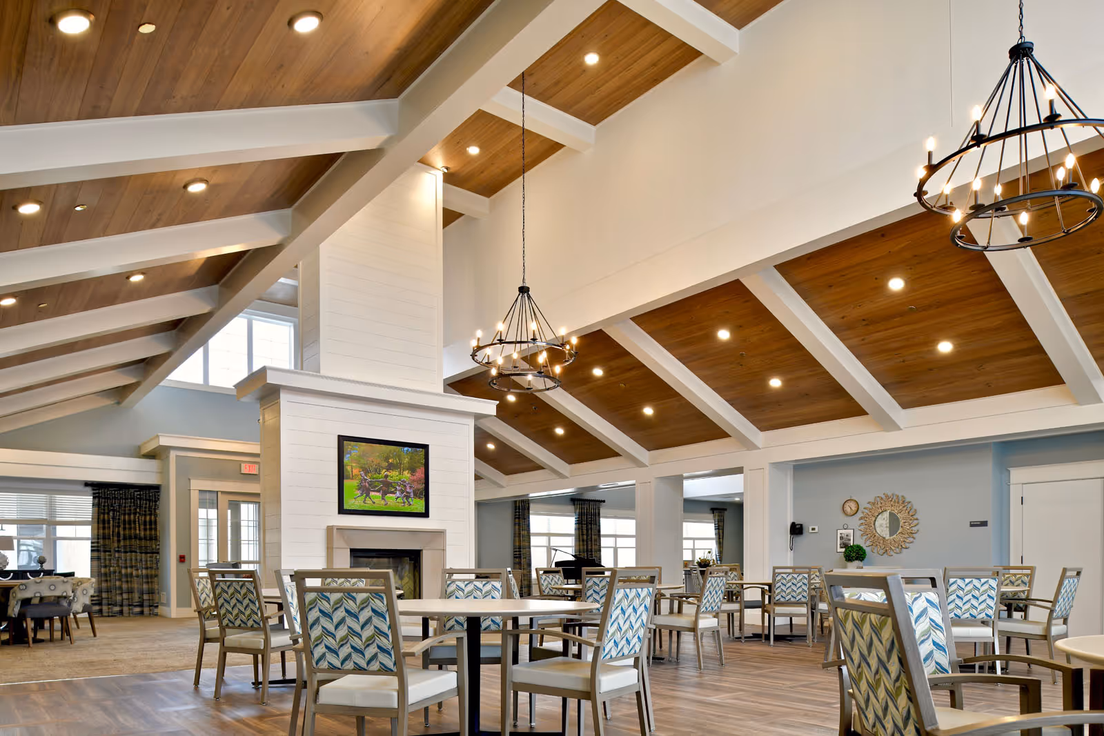 Spacious dining and lounge area with vaulted wood-paneled ceiling, chandeliers, a central fireplace with TV, and multiple tables and chairs.