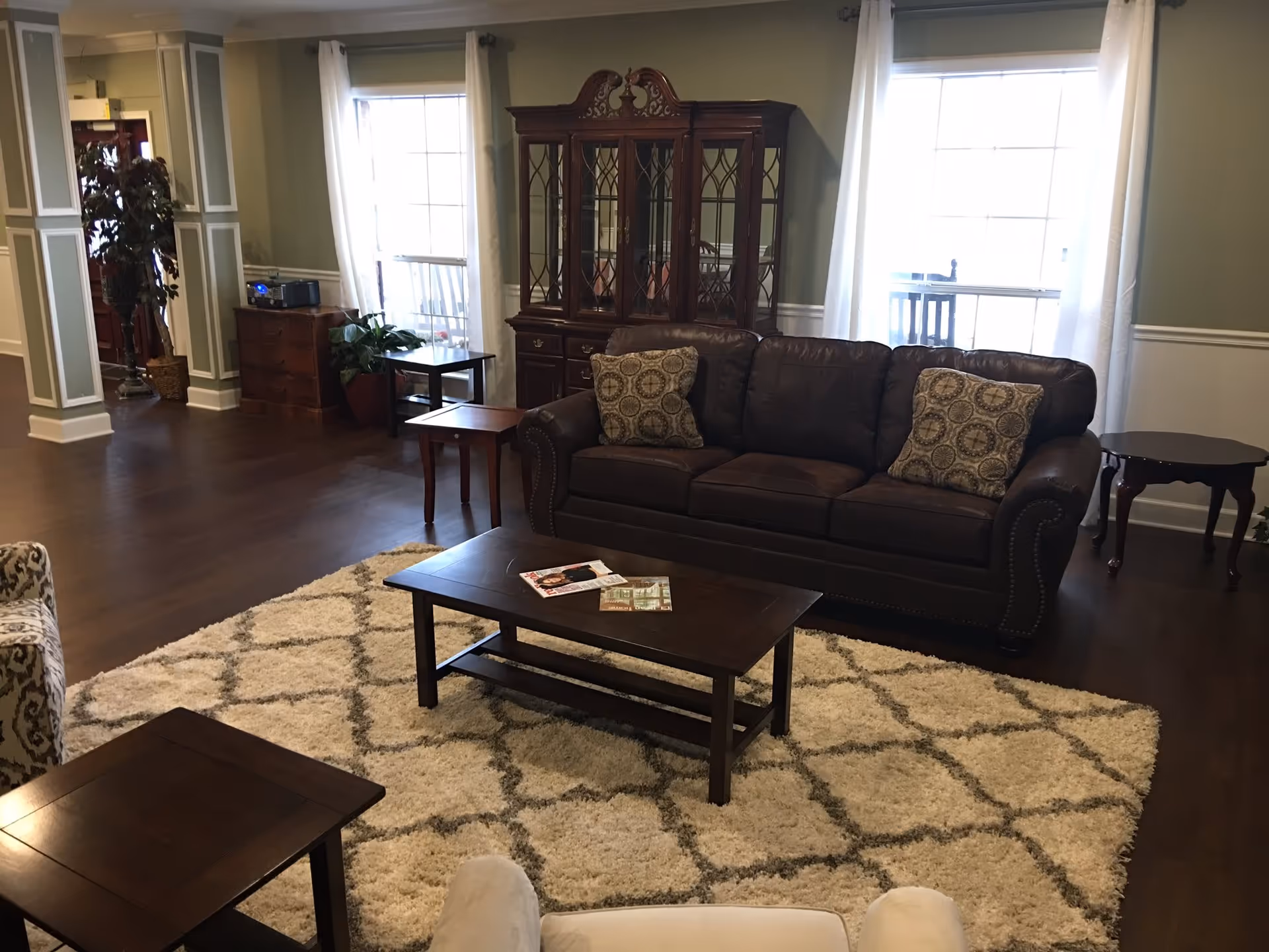 A cozy living room with a dark brown leather sofa adorned with patterned cushions, a wooden coffee table with magazines on it, a cream and gray patterned rug, and a wooden china cabinet against the wall. The room has two large windows with white curtains, wooden flooring, and several side tables. There is also a potted plant near the entrance.