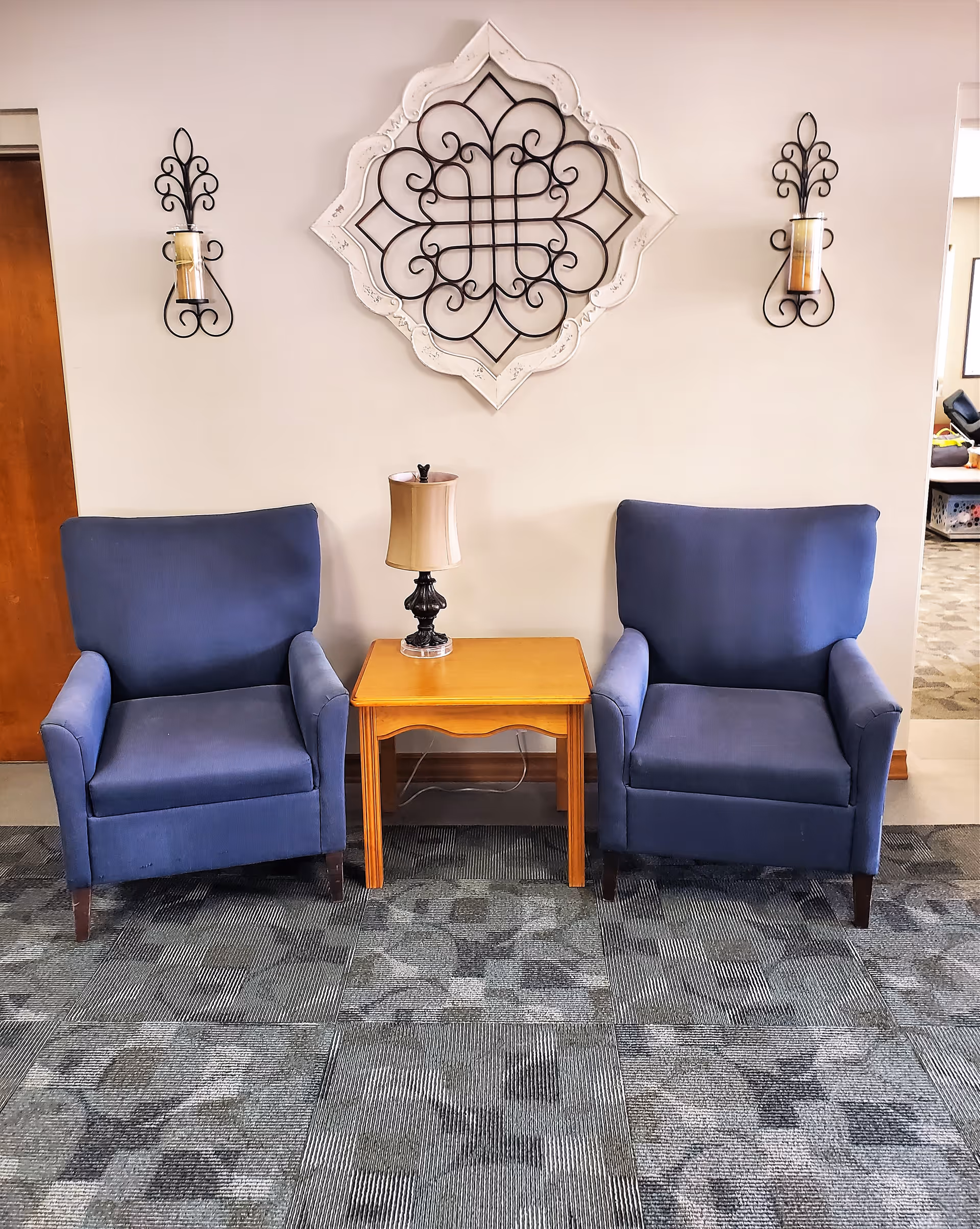 Two blue armchairs flank a wooden side table with a lamp beneath decorative wall art and candle sconces.
