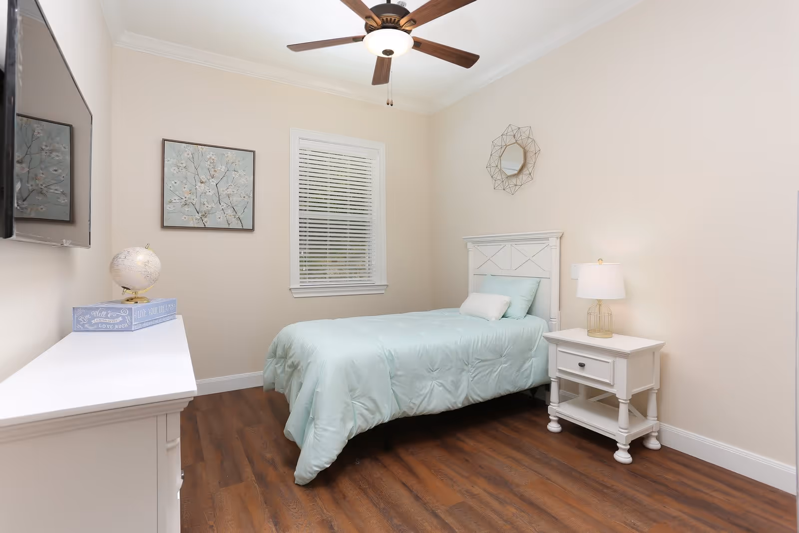 A neatly arranged bedroom with a single bed covered in a light blue comforter and pillows. The room features a white nightstand with a lamp, a white dresser with a decorative globe and a box on top, a ceiling fan with a light, a window with blinds, and two pieces of wall art including a floral painting and a geometric mirror.