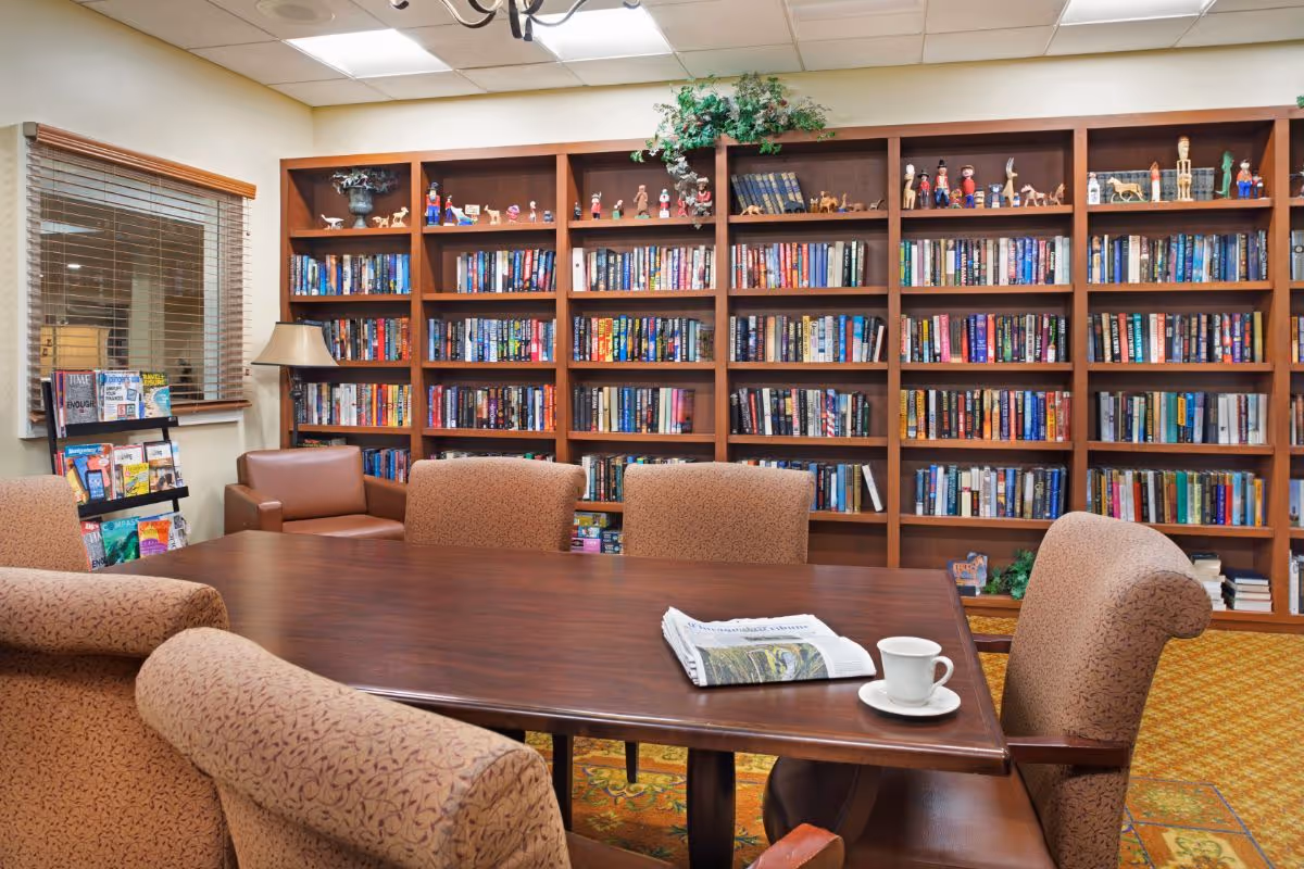 A cozy community library/reading room with a large wooden table surrounded by upholstered chairs, bookshelves filled with books, and a newspaper and coffee cup on the table.