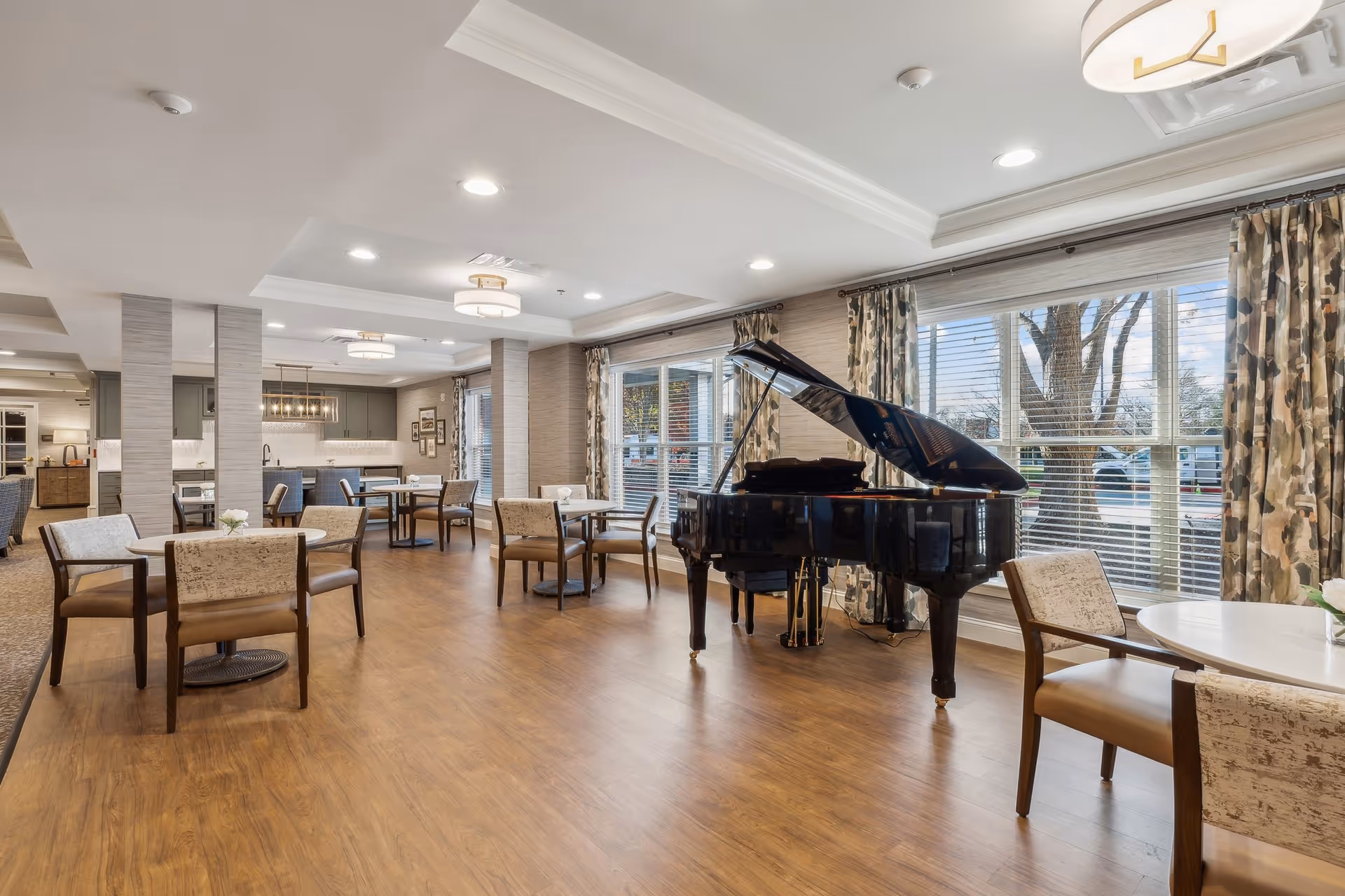 A spacious, well-lit common area in a senior living facility featuring several round tables with chairs, a black grand piano near large windows with patterned curtains, and a kitchenette area in the background with modern cabinetry and pendant lighting.