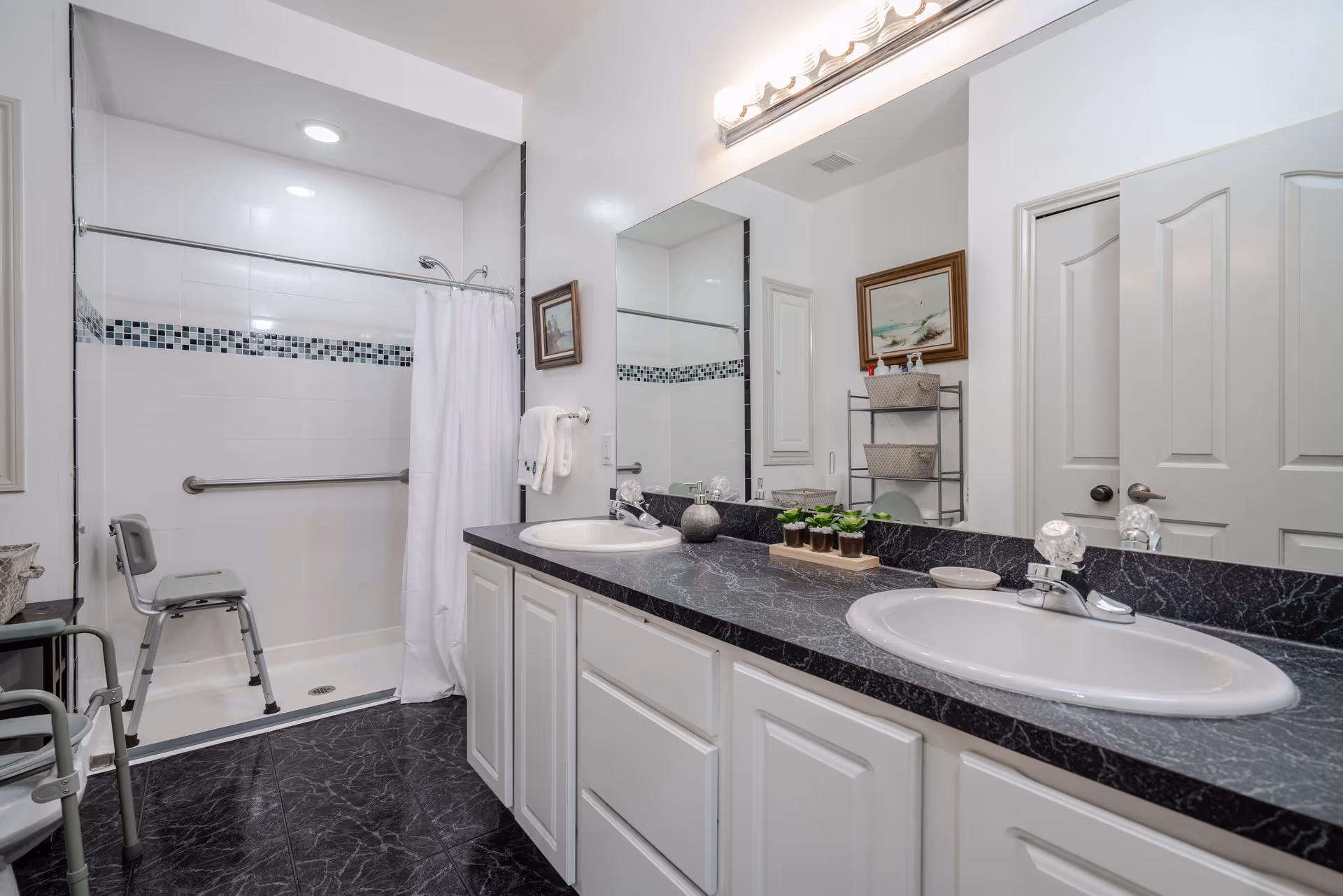 A clean and modern bathroom featuring a double sink vanity with black marble countertop and white cabinets. There is a large mirror above the sinks with a light fixture. To the left, there is a walk-in shower with white tiles and a decorative blue and black tile border, equipped with a shower chair and grab bars for accessibility. The floor has dark marble tiles, and there is a metal shelving unit with baskets and a framed picture on the wall.