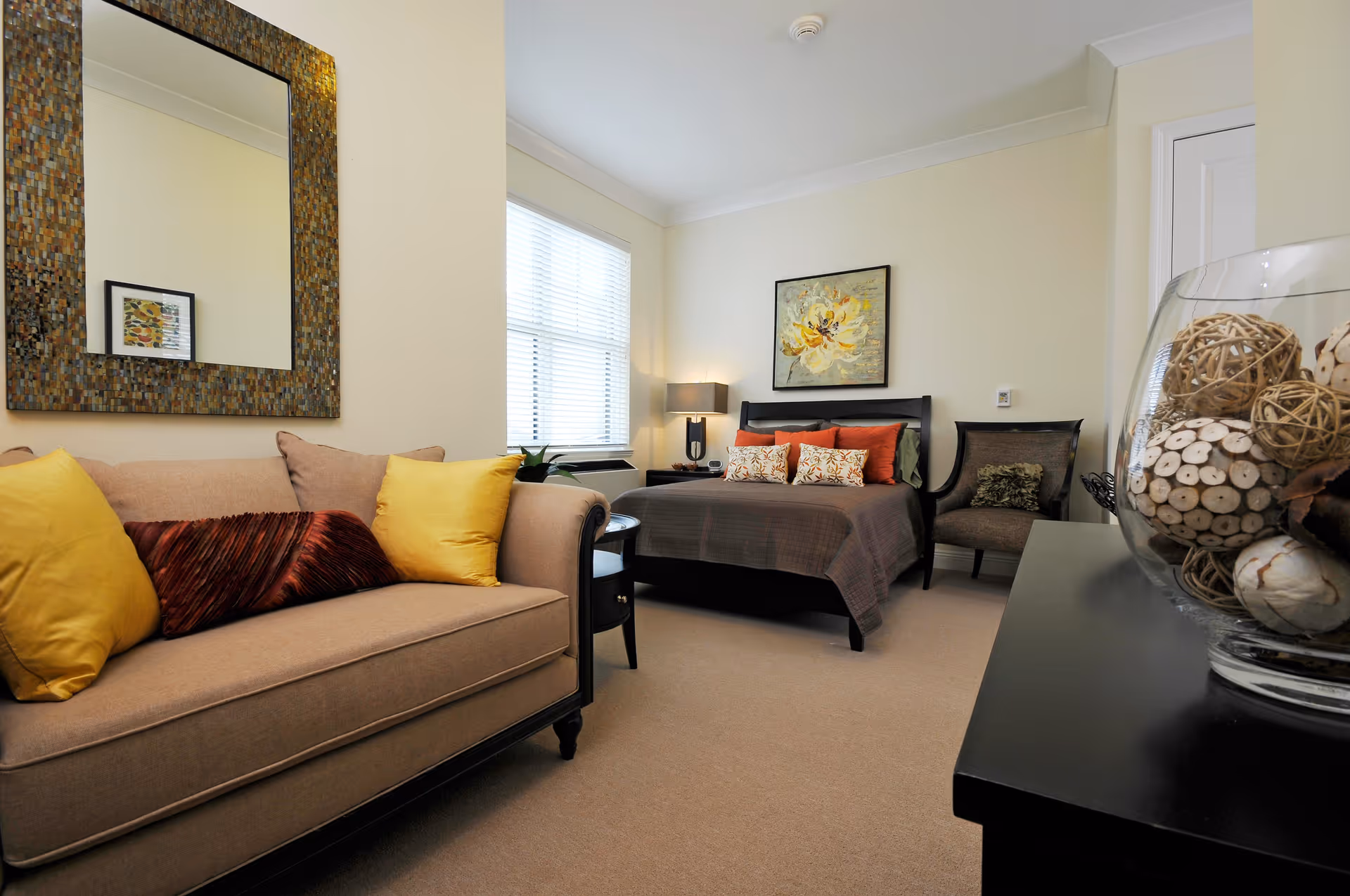 A cozy bedroom featuring a bed with multiple pillows and a brown bedspread, a beige sofa with yellow and brown cushions, a large decorative mirror on the wall, a side table with a lamp, a framed floral painting above the bed, a chair in the corner, and a glass bowl filled with decorative natural elements on a black dresser.