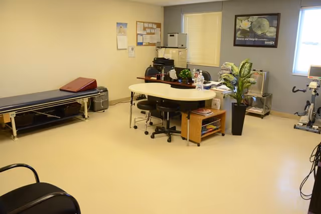 A bright interior therapy room with a central oval table and rolling stools, an exam/treatment bench, exercise equipment, filing cabinets and a potted plant.