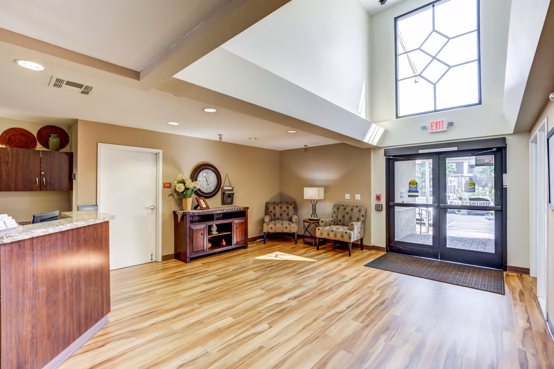 A bright and spacious senior living facility lobby with wood flooring, a reception desk on the left, two patterned armchairs with a small table and lamp in the corner, a wooden cabinet with decorative items and a wall clock, and large glass double doors leading outside under a high ceiling with a large window.