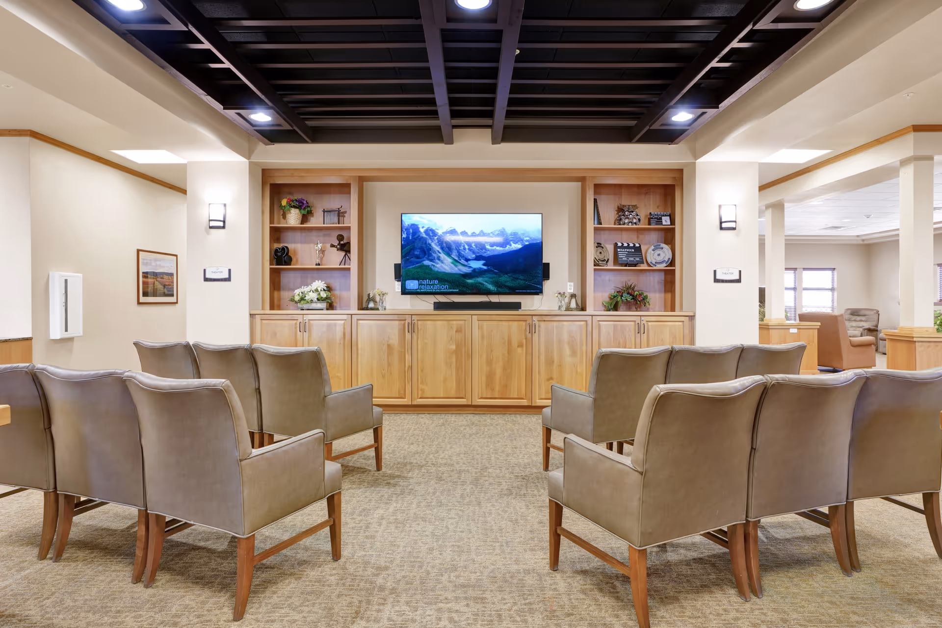 A cozy common area with two rows of beige armchairs facing a wall-mounted flat-screen TV. The TV is set within a wooden built-in cabinet with shelves displaying decorative items and plants. The room has beige walls, carpeted flooring, and recessed lighting in a dark ceiling grid. Adjacent areas with additional seating are visible in the background.