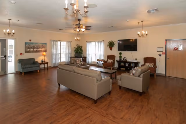 Spacious furnished common living room with sofas, armchairs, a TV, coffee table and chandeliers in a senior care facility.