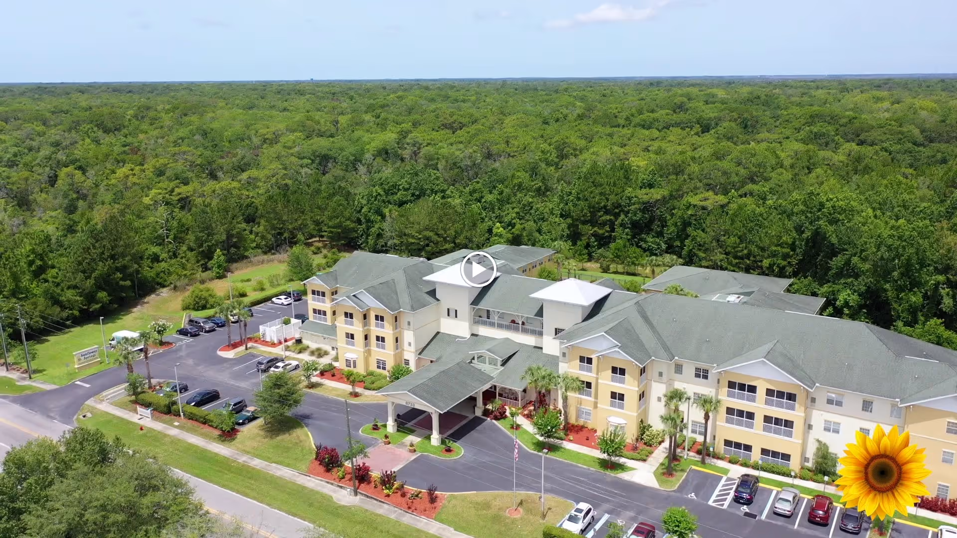 Aerial view of Sunflower Springs senior living facility surrounded by dense green forest. The building is a large, multi-story structure with a covered entrance and several parked cars in the parking lot. The sky is clear and blue.