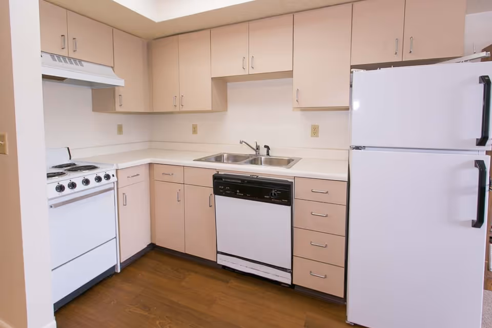 A compact kitchen with beige cabinets, a white electric stove with four burners, a white dishwasher, a double stainless steel sink, and a white refrigerator. The kitchen has a wooden floor and white countertops.
