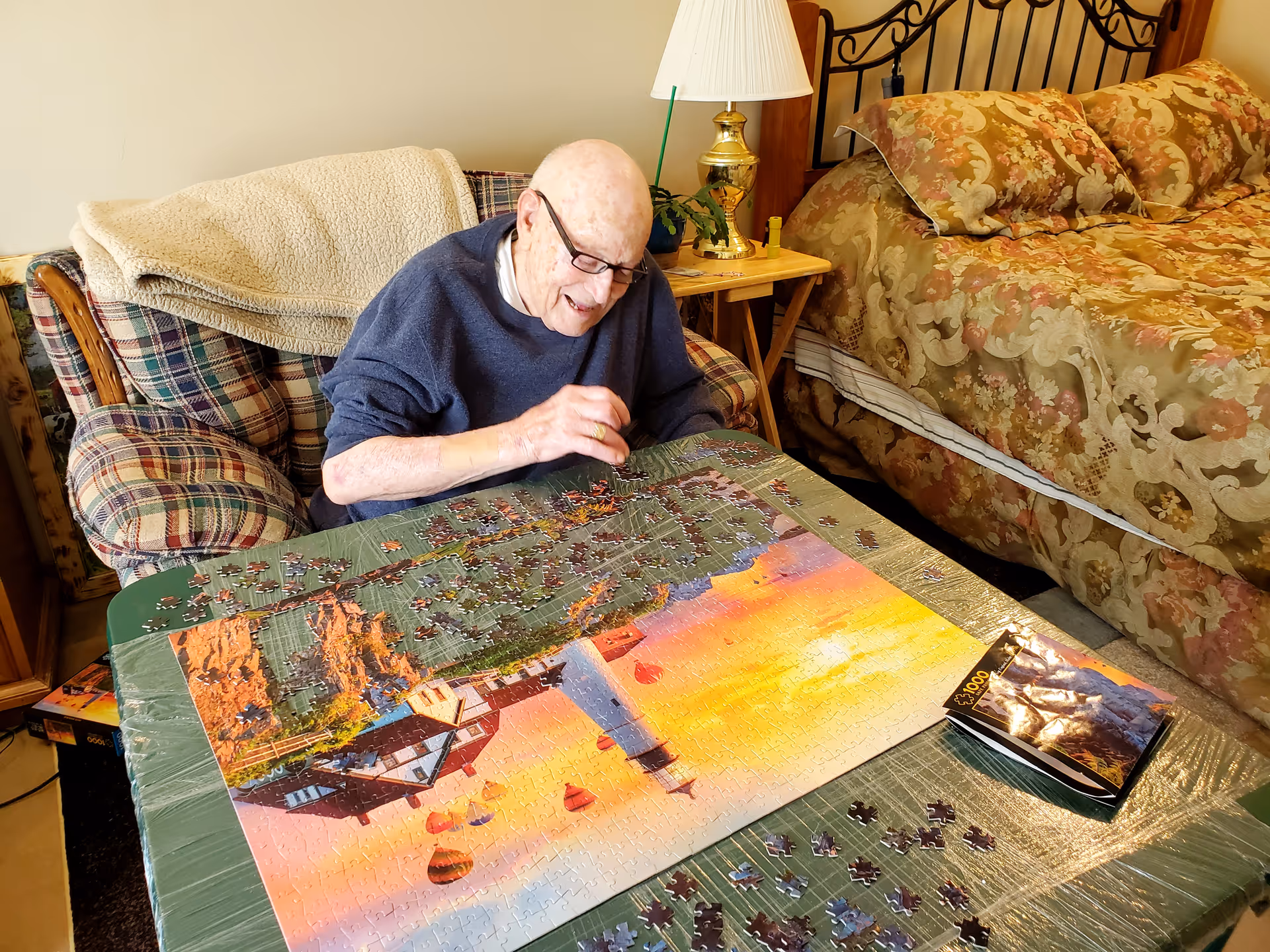 An elderly man wearing glasses and a dark sweater is sitting at a table working on a large jigsaw puzzle featuring a lighthouse and a sunset. He is in a cozy room with a plaid armchair, a wooden side table with a lamp, and a bed with floral bedding in the background.