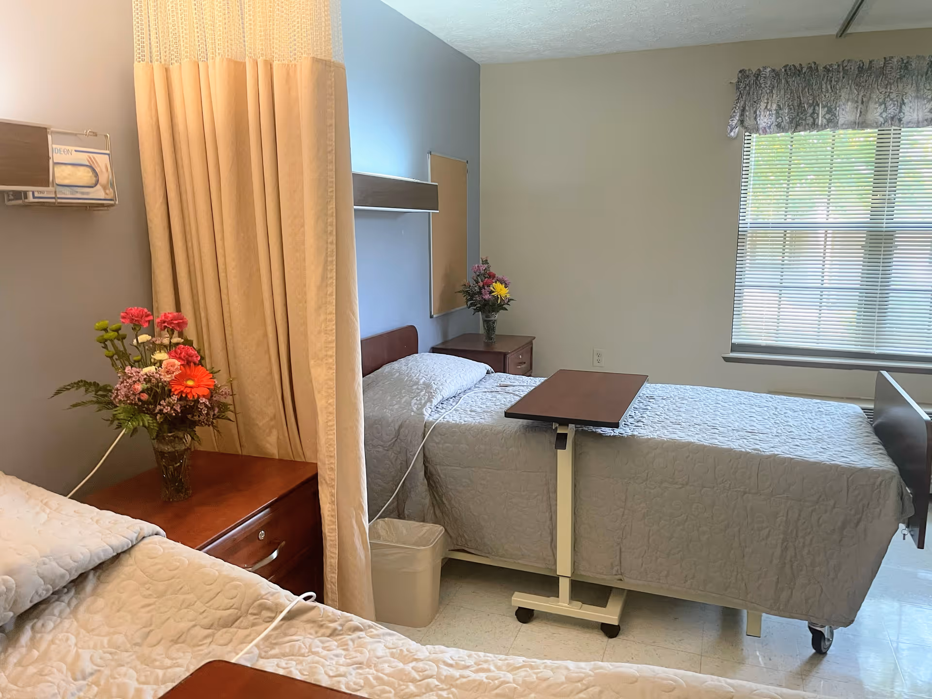 A nursing facility room with two hospital beds separated by a beige privacy curtain. Each bed has a light-colored quilted bedspread, and there are bedside tables with flower vases. A window with blinds and a floral valance lets in natural light.