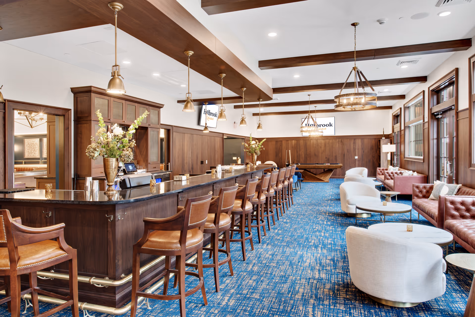 A spacious and elegant lounge area featuring a long wooden bar with leather bar stools, pendant lights hanging from the ceiling, and a blue patterned carpet. The room also includes comfortable white armchairs and brown tufted sofas arranged around small round tables. Large windows line one side of the room, allowing natural light to enter. Two screens on the far wall display the text 'Holbrook Woodstock'.
