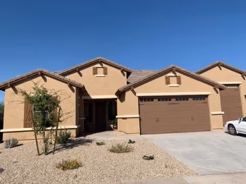 Exterior view of a single-story beige stucco house with a brown tiled roof, a two-car garage, a driveway, and a small front yard with desert landscaping including gravel and sparse plants under a clear blue sky.