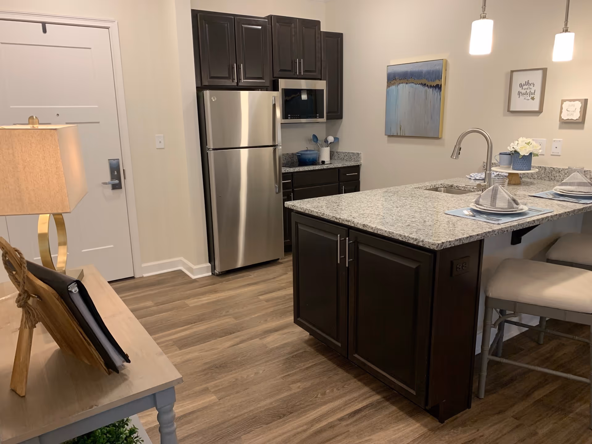 Modern kitchen area with dark wood cabinets, stainless steel refrigerator and microwave, granite countertop island with two place settings, and pendant lights hanging above. A lamp and some decor are visible on a small table near the entrance door.