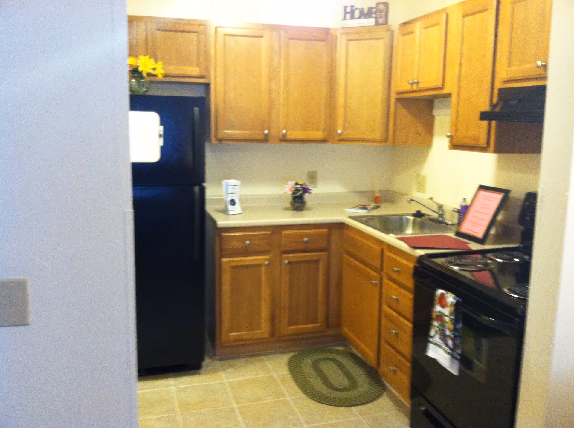 Small kitchen with wooden cabinets, a black refrigerator, black stove, stainless steel sink, and beige countertops. There is a small green rug on the tiled floor and decorative items on the counter and above the cabinets.