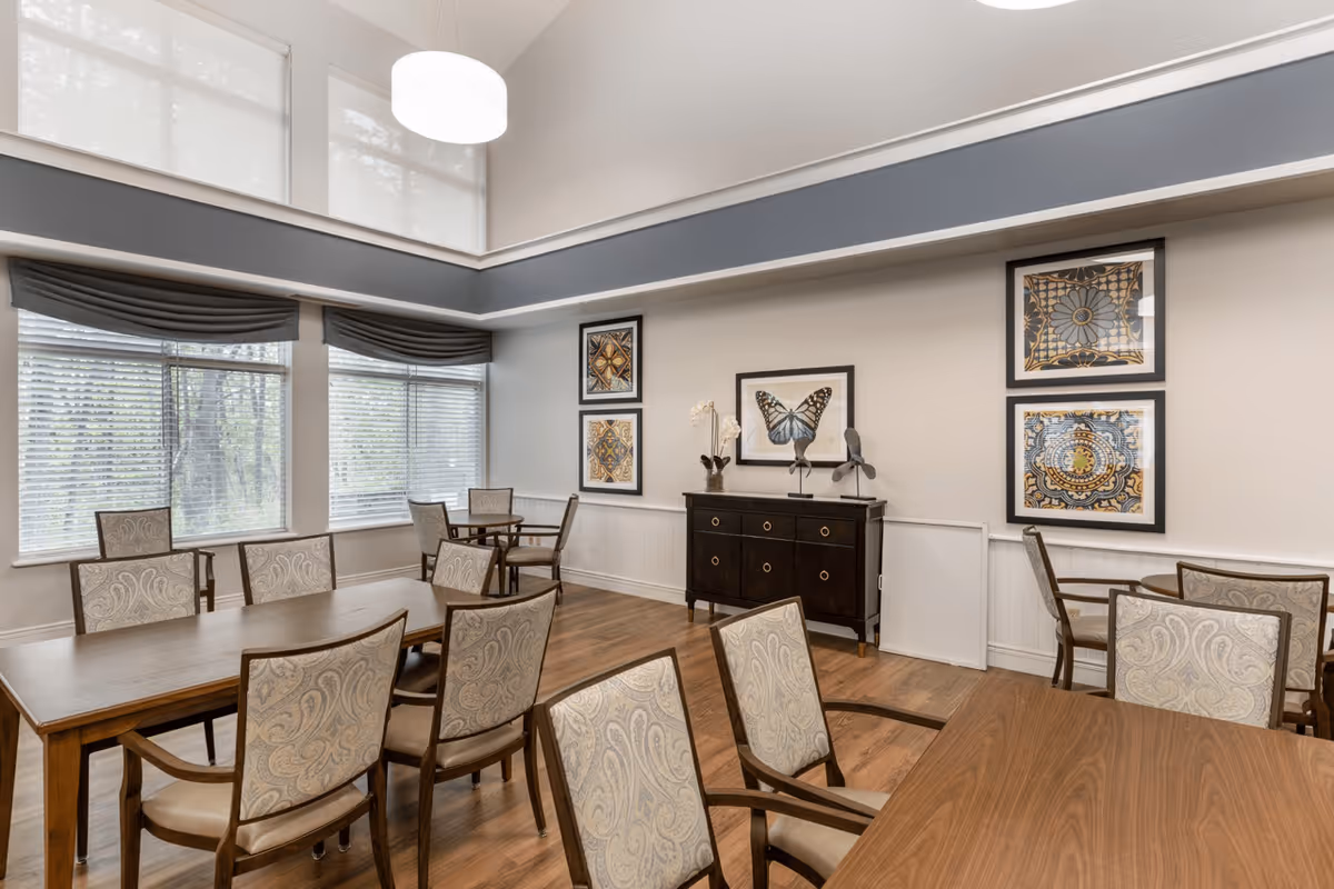 A bright dining room with multiple wooden tables and upholstered chairs arranged neatly. Large windows with blinds and valances allow natural light to fill the space. The walls are decorated with framed artwork including a butterfly and intricate patterns. A dark wooden sideboard with decorative items is positioned against one wall. The room has a high ceiling with a modern pendant light fixture.