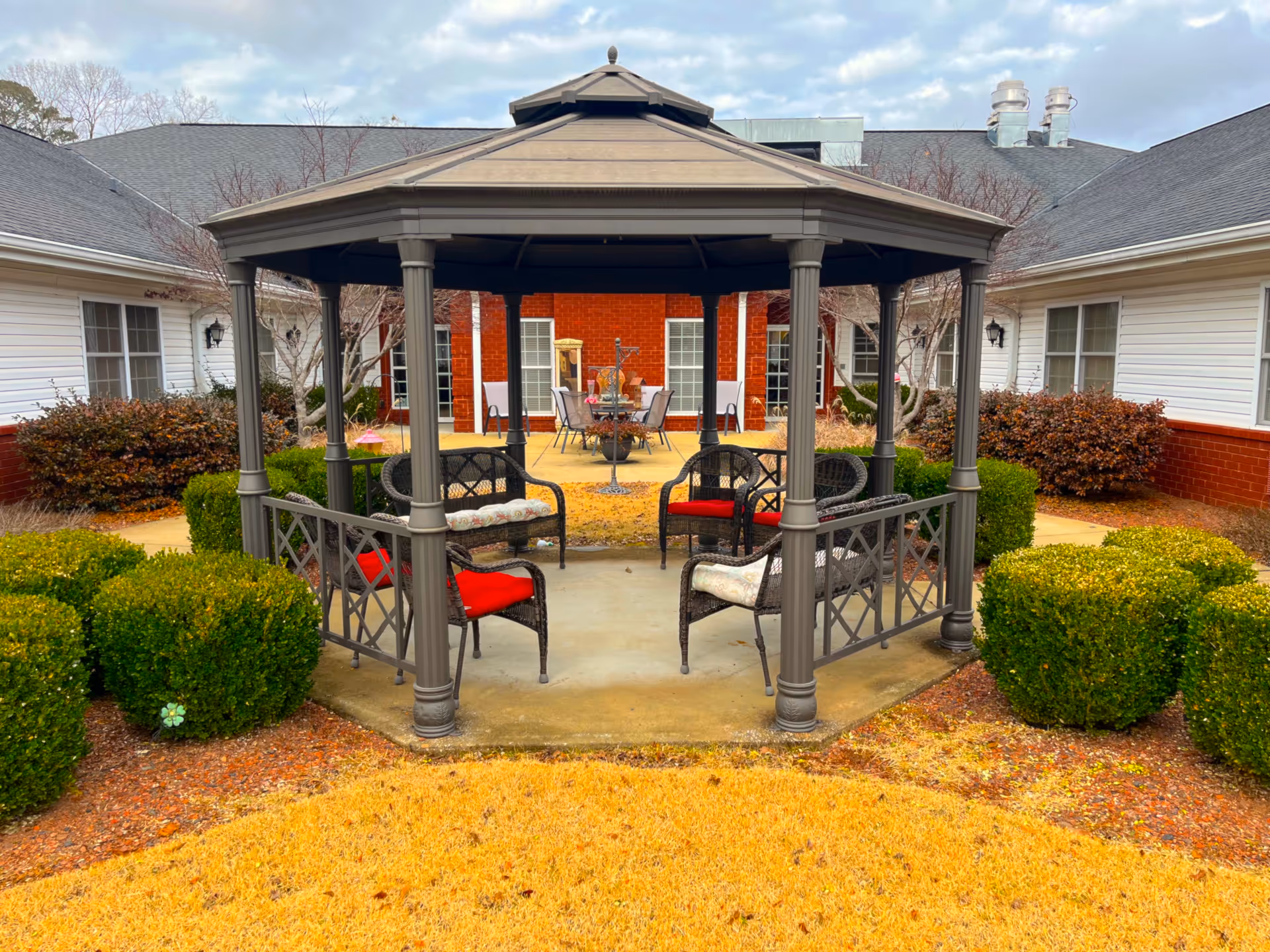 Outdoor gazebo with four cushioned chairs arranged inside, surrounded by neatly trimmed bushes and yellow grass. The gazebo is located in a courtyard area between two white buildings with red brick lower walls and multiple windows. In the background, there is a patio area with a table and chairs.