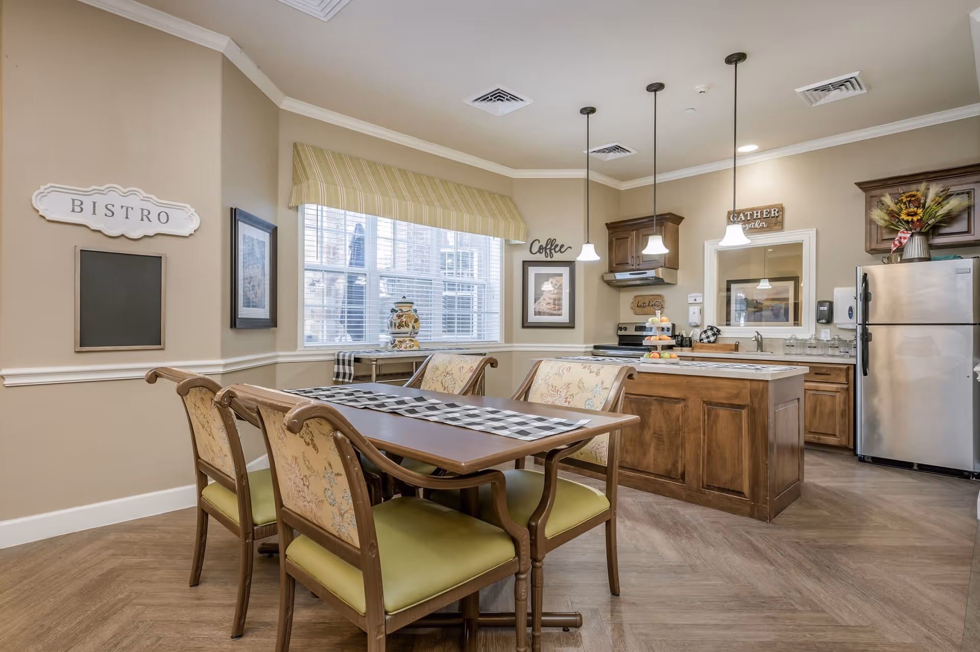 A cozy dining area and kitchen in a senior living facility. The dining area features a wooden table with four cushioned chairs, a checkered table runner, and a large window with a striped valance. The kitchen has wooden cabinets, a stainless steel refrigerator, a stove, a kitchen island with pendant lights above, and decorative signs on the walls including 'Bistro', 'Coffee', and 'Gather together'.