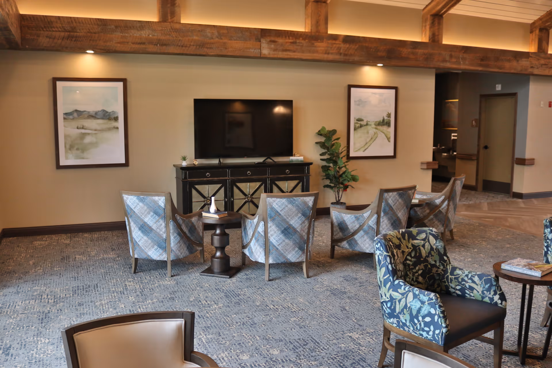 A cozy living room area with a large flat-screen TV mounted on a black cabinet. In front of the TV are four blue plaid armchairs arranged in a row, with a small round table between the two middle chairs. To the right, there is a blue floral armchair next to a small round wooden table with books on it. The room has beige walls adorned with two framed landscape paintings and a potted plant near the TV cabinet. The ceiling features exposed wooden beams with recessed lighting, and the floor is covered with a patterned carpet.