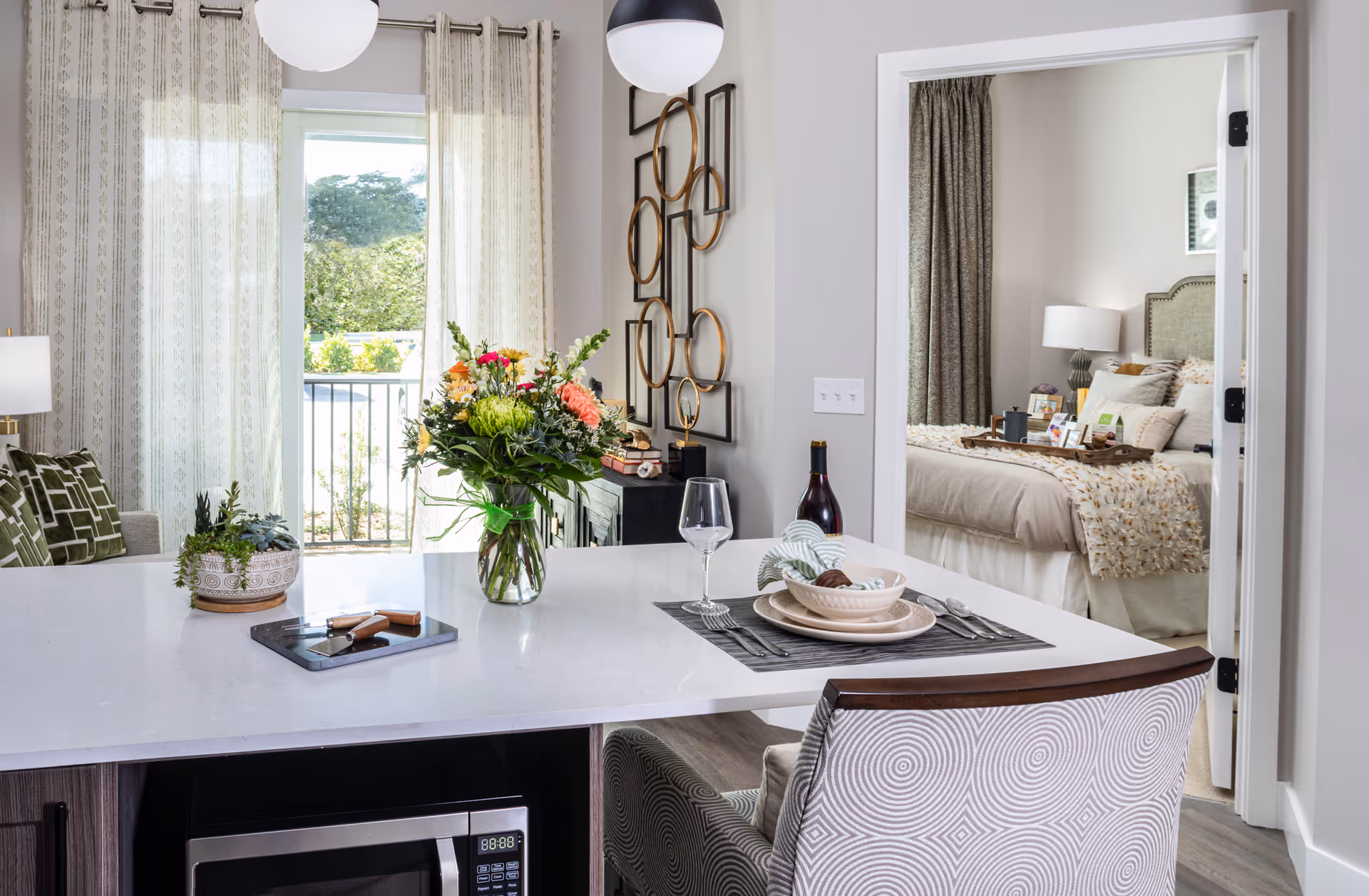 A modern interior space featuring a white countertop with a vase of colorful flowers, a wine bottle, and a place setting with plates, a bowl, and utensils. Behind the countertop is a living area with patterned curtains and a decorative wall piece. Through an open doorway, a bedroom is visible with a neatly made bed, a lamp, and framed artwork on the wall.