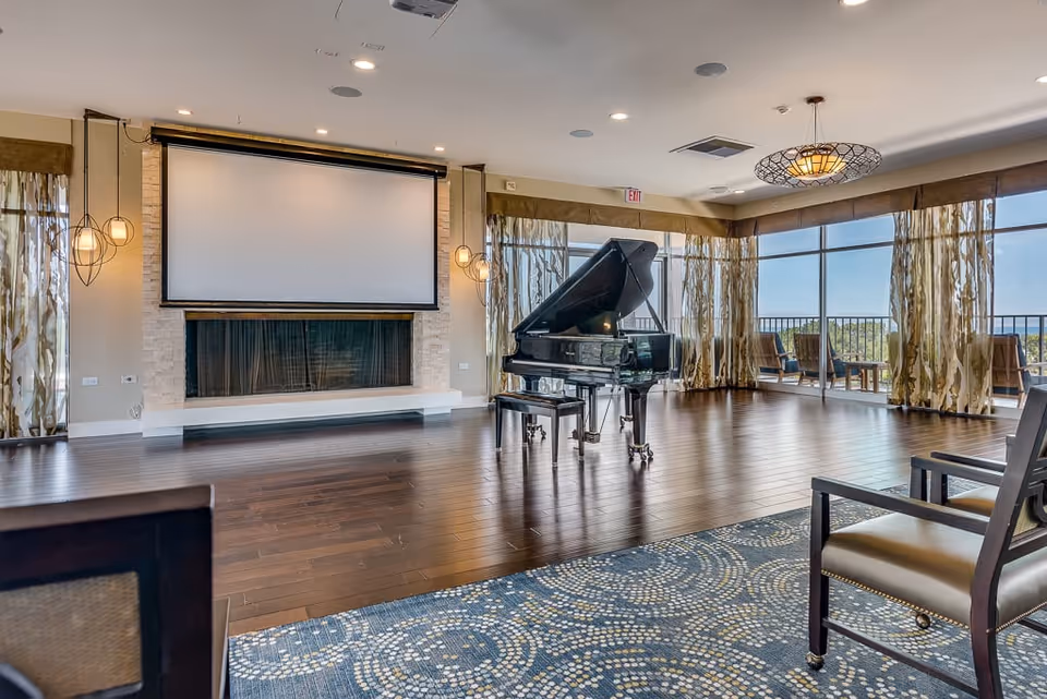 A spacious senior living common area with a grand piano in the center, hardwood floors, large windows with patterned curtains allowing natural light, a large projector screen above a fireplace, and several chairs arranged around the room.