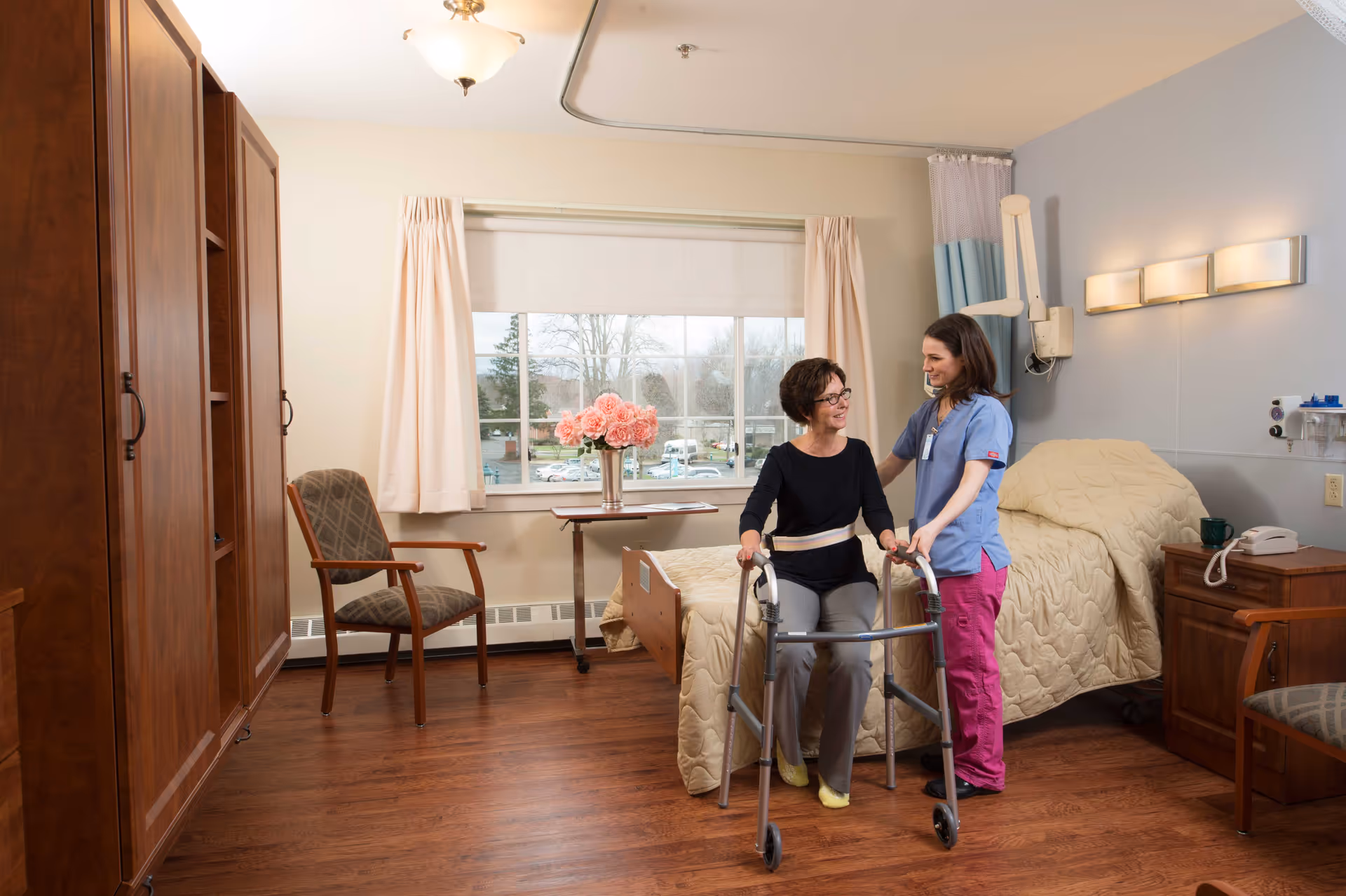 A senior woman using a walker is assisted by a female caregiver in a well-lit bedroom with wooden furniture, a bed with a beige quilt, a window with curtains, and a vase of pink flowers on a table.