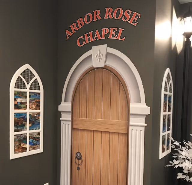 Interior wall with a decorative faux wooden chapel door labeled 'Arbor Rose Chapel' above it, flanked by two white-framed stained glass window decorations and a black lamp post style light fixture on the right side.
