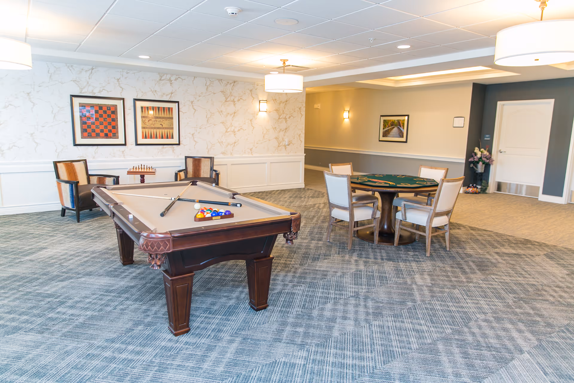 A spacious game room in a senior living facility featuring a pool table with cues and balls arranged for play, a round card table with four chairs, two armchairs with a chessboard between them, framed artwork on the walls, and soft overhead lighting.