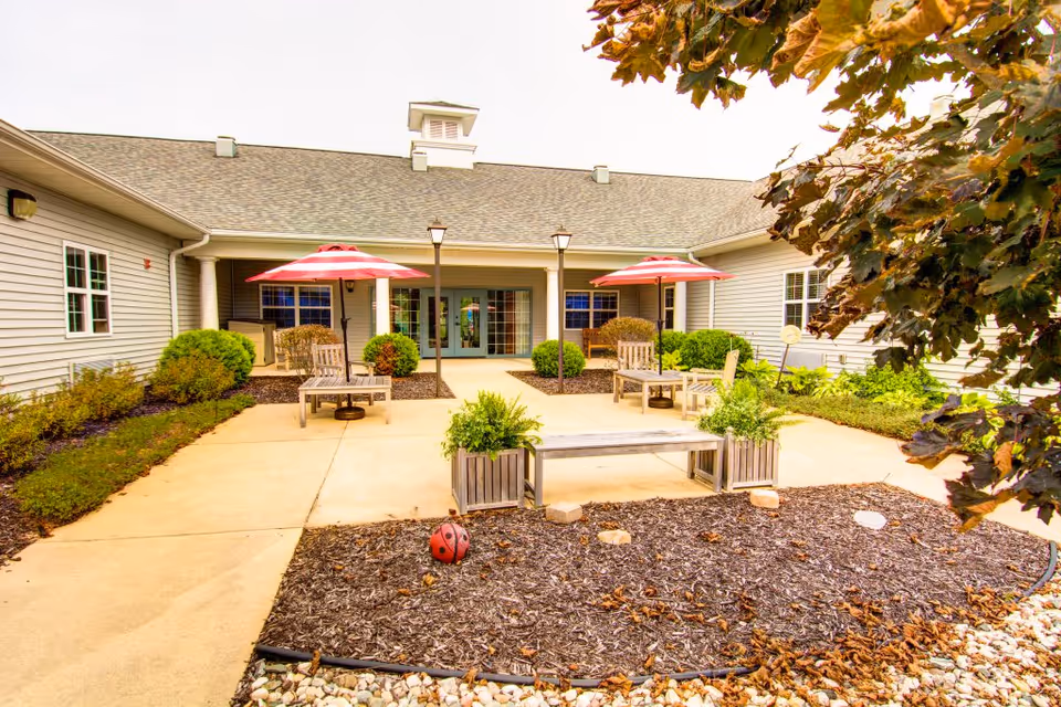 Outdoor courtyard area of a senior living facility with a concrete walkway, wooden benches, chairs, and tables shaded by red umbrellas. The courtyard is surrounded by bushes and plants, with a building featuring multiple windows and a covered entrance in the background. Trees with autumn leaves frame the right side of the image.