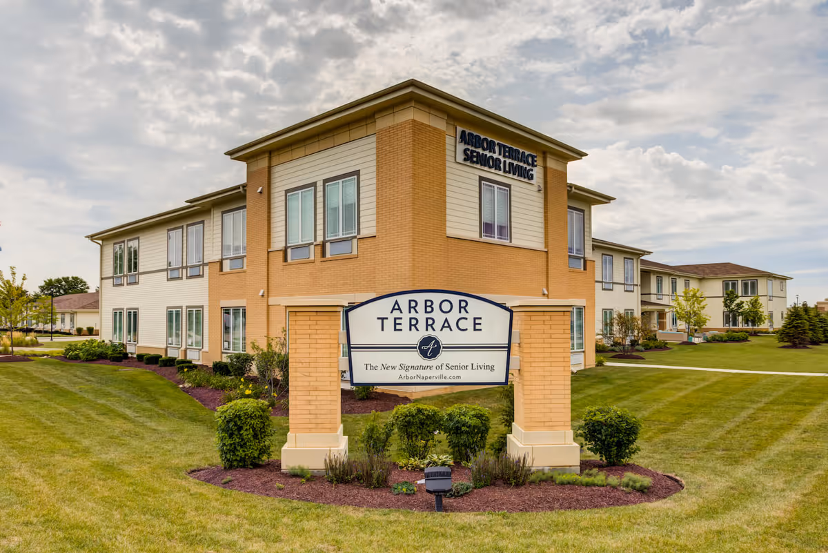 Exterior view of Arbor Terrace Naperville senior living facility showing a two-story building with beige and light brown walls, multiple windows, and a landscaped lawn with bushes and trees under a cloudy sky. A large sign in front reads 'Arbor Terrace - The New Signature of Senior Living - ArborNaperville.com'.